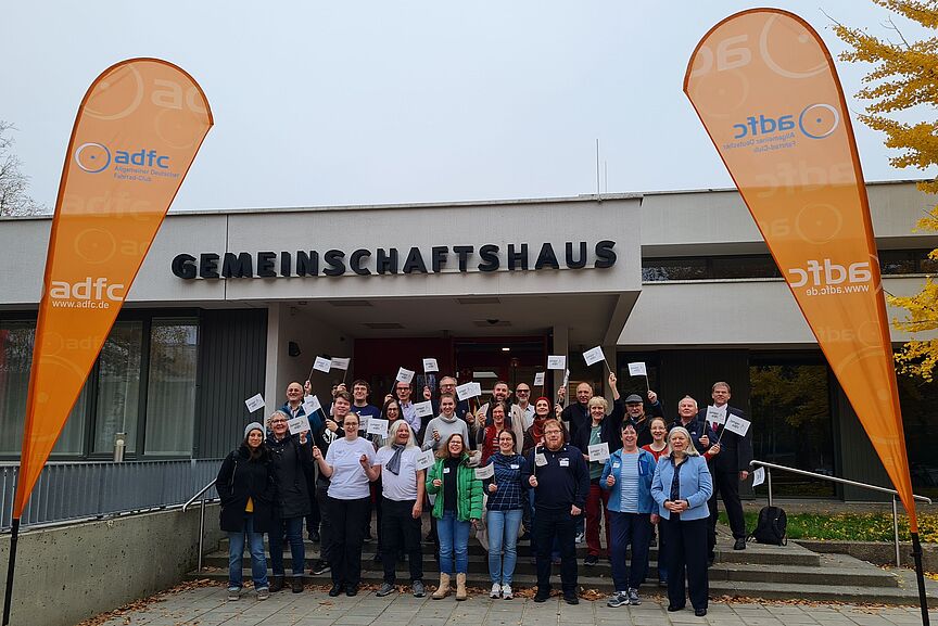 2024_BHV_Delegiertengruppe_IK Eine große Gruppe Menschen steht vor einem Gebäude. Die Menschen tragen überwiegend Winterkleidung. Einige von Ihnen schwenken Fahnen, auf denen das Logo des Jungen ADFC zu sehen ist. Auf dem Gebäude steht "Gemeinschaftshaus". Am linken und rechten Bildrand sind orange Flaggen aufgestellt, die das Logo des ADFC zeigen.