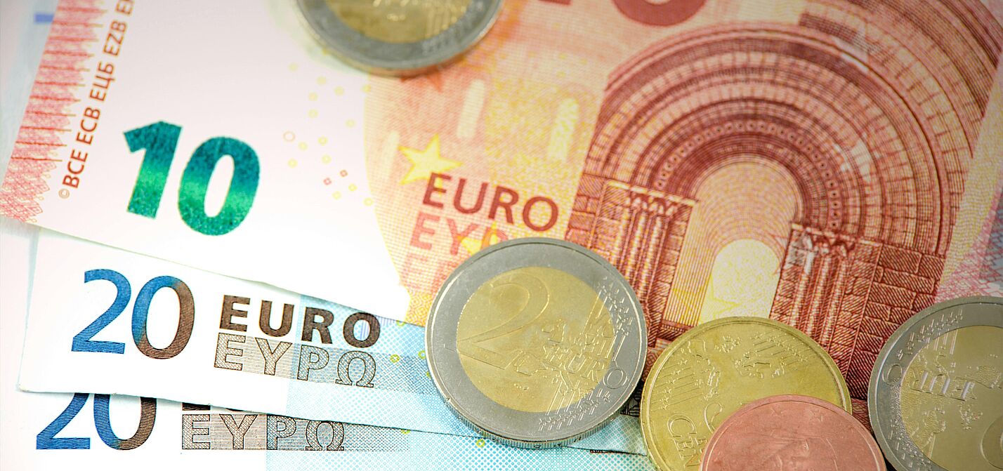 Es sind mehrere Geldscheine zu sehen. Ein Zehn-Euro-Schein liegt auf mehreren Zwanzig-Euro- und einem Fünf-Euro-Schein. Außerdem liegen einige Münzen auf den Scheinen verstreut.